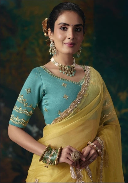Yellow tissue silk saree with ornate gold border, draped over a teal blouse with embroidered sleeves